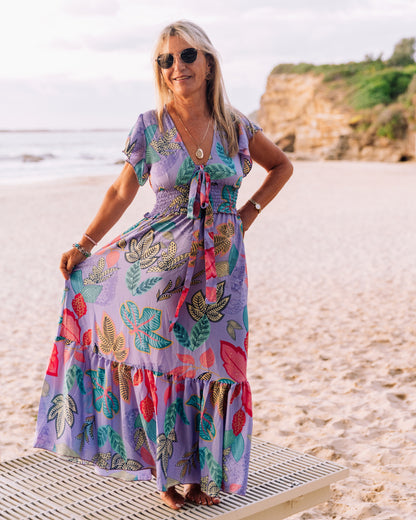 woman at the beach wearing a floral leaf print flutter sleeve with tie front maxi dress in purple